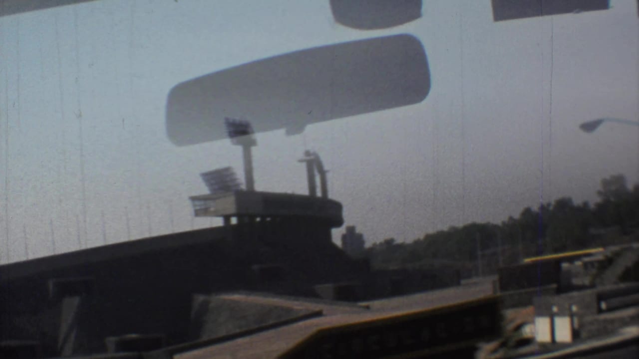 Drive-by view of a concrete stadium with floodlights seen through a car windshield.