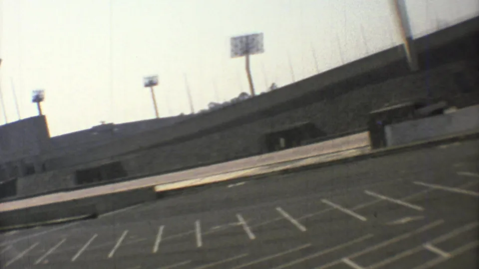 1973 Mexico City arena exterior seen from a low handheld angle, with an empty striped parking lot and floodlight towers above the concrete structure.