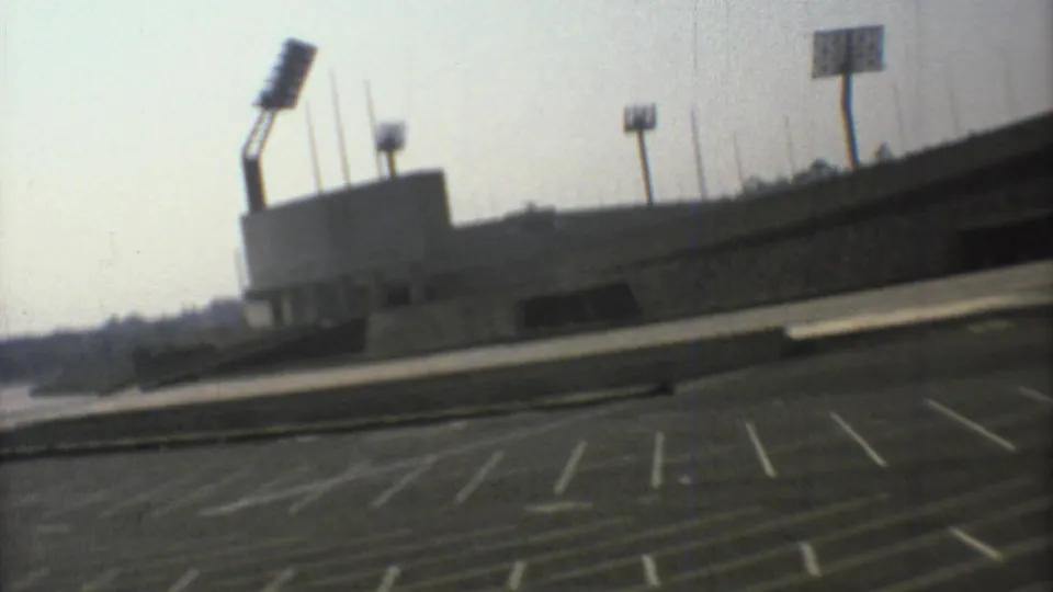 Handheld 8mm view of the Mexico City sports arena exterior, showing floodlight poles, concrete walls, and a mostly empty marked parking area.