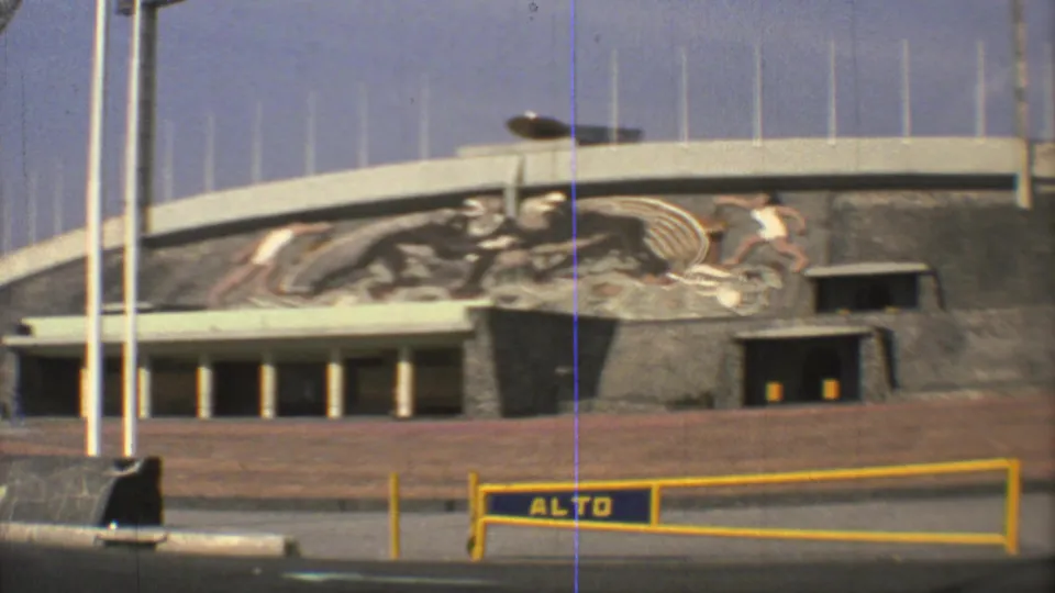 Mexico City sports arena facade with a large mural of athletes, tall flagpoles, entrance doors, and a yellow barrier marked with visible lettering.