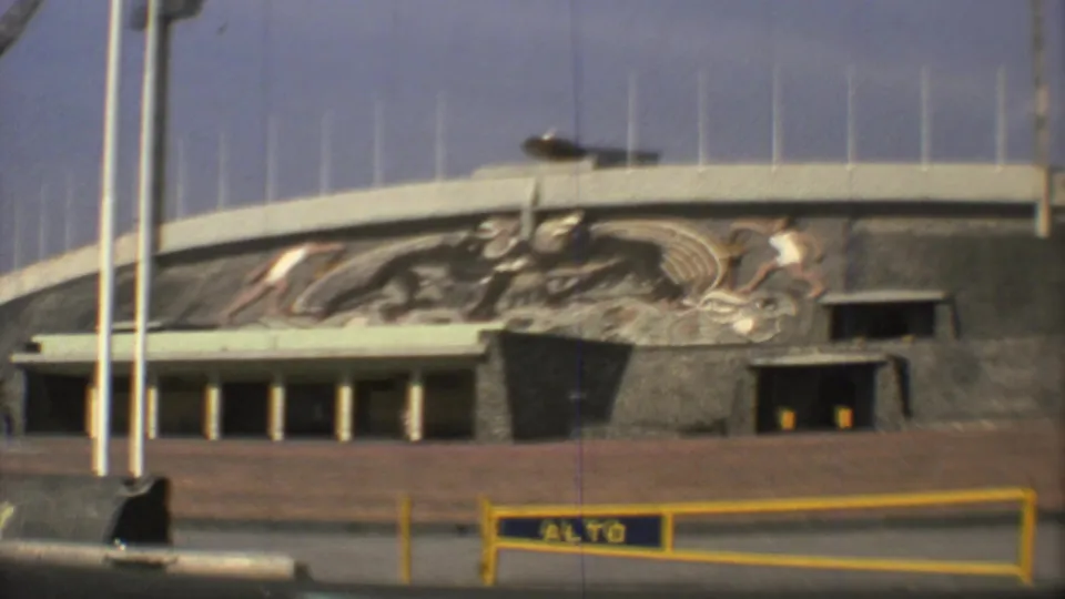 1973 Mexico City 8mm frame showing a modernist building facade with a large sports mural, flagpoles, and an ALTO barrier in front.