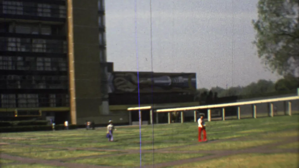 1973 Mexico City 8mm frame showing people walking across green grounds near a tall blocky building and a long covered walkway.