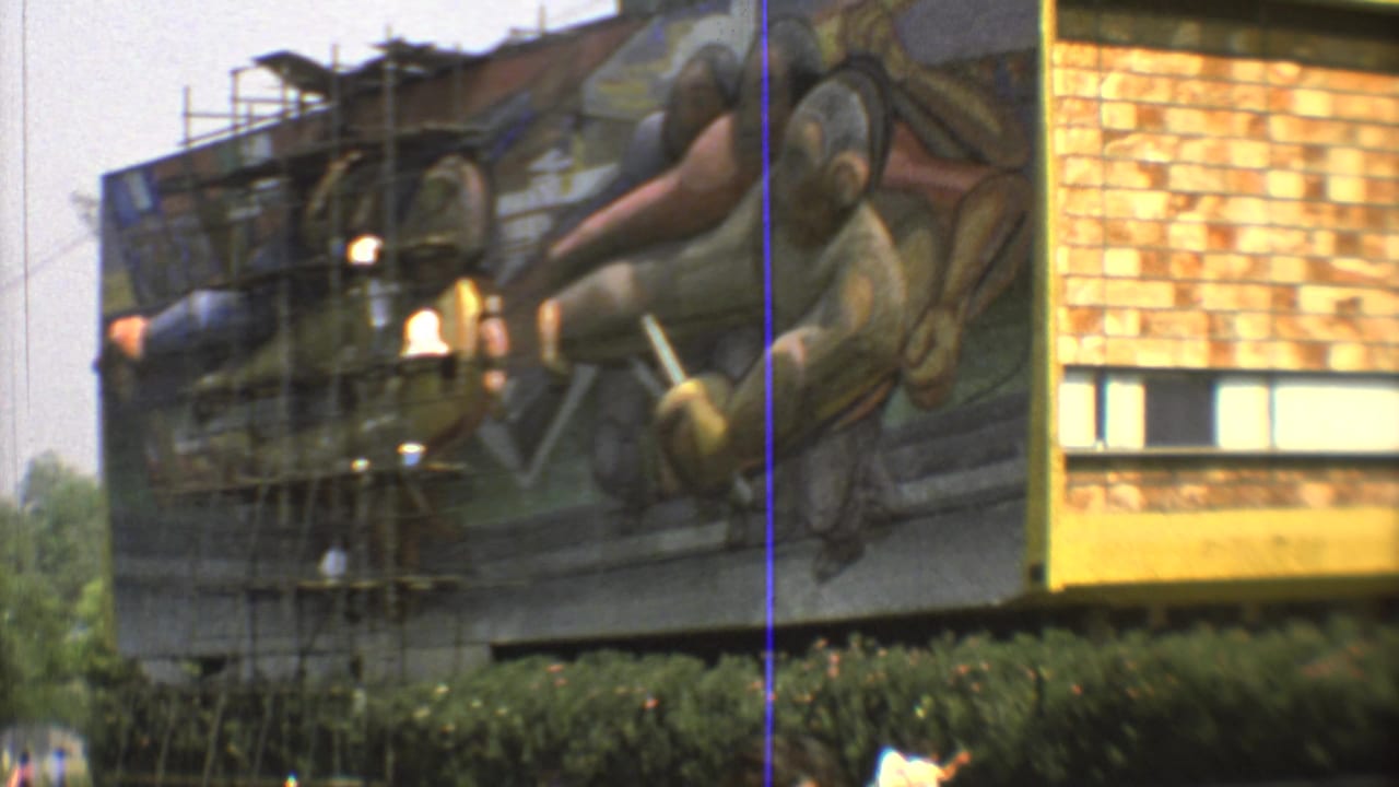 1973 Mexico City mural facade with scaffolding over painted figures, a brick wall section, hedge, and distant people below.