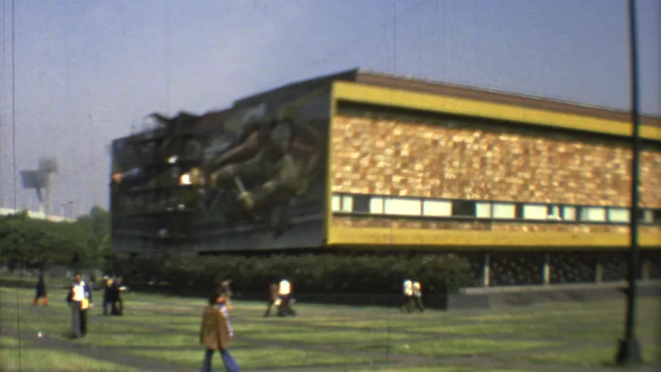 Wide 1973 Mexico City view of a modernist building with yellow trim, a mural at one end, and pedestrians crossing the lawn.