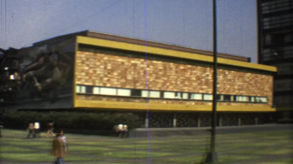 1973 Mexico City frame showing a long yellow-trimmed facade, a partial mural at left, pedestrians, and a lamppost in the foreground.