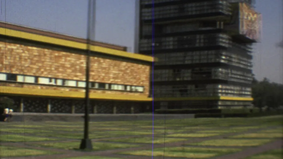 End of the 1973 Mexico City pan showing the modernist building beside a taller tower, with lawn, paths, and lampposts in view.