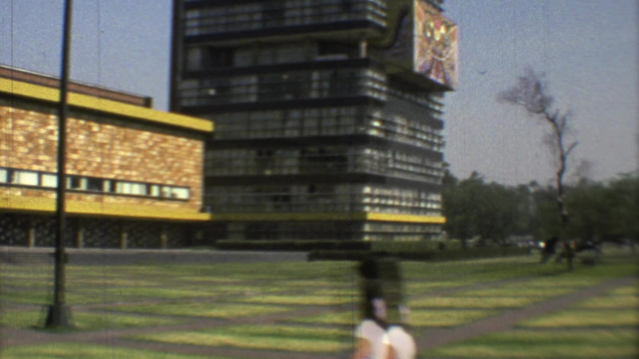 1973 Mexico City park view with a dark modernist building, yellow-trimmed structure, patterned grass, and a passing pedestrian.