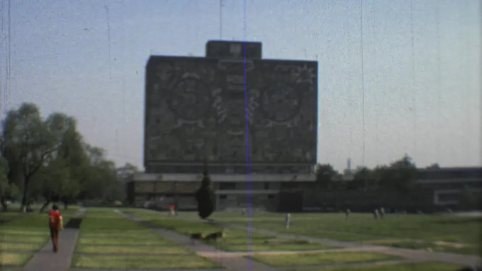 1973 Mexico City 8mm frame with the carved stone monument, diagonal park paths, a red-shirted walker, a dog, trees, and distant buildings.