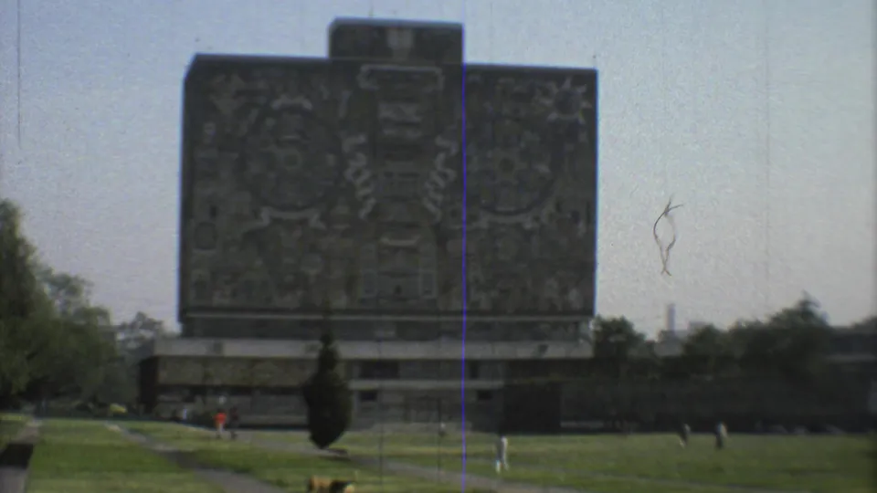 1973 Mexico City frame showing a closer view of the carved stone monument, surrounding lawns, trees, distant pedestrians, and a low structure.