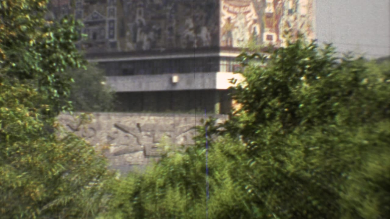 1973 Mexico City 8mm frame showing dense greenery partially obscuring tall mural-like building faces and a stone or concrete relief wall.