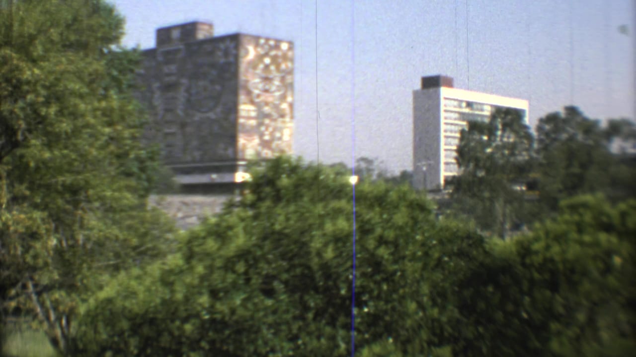 Final wide 1973 Mexico City frame with lush trees, a tall painted building surface, a pale tower, and a relief wall partly visible below.