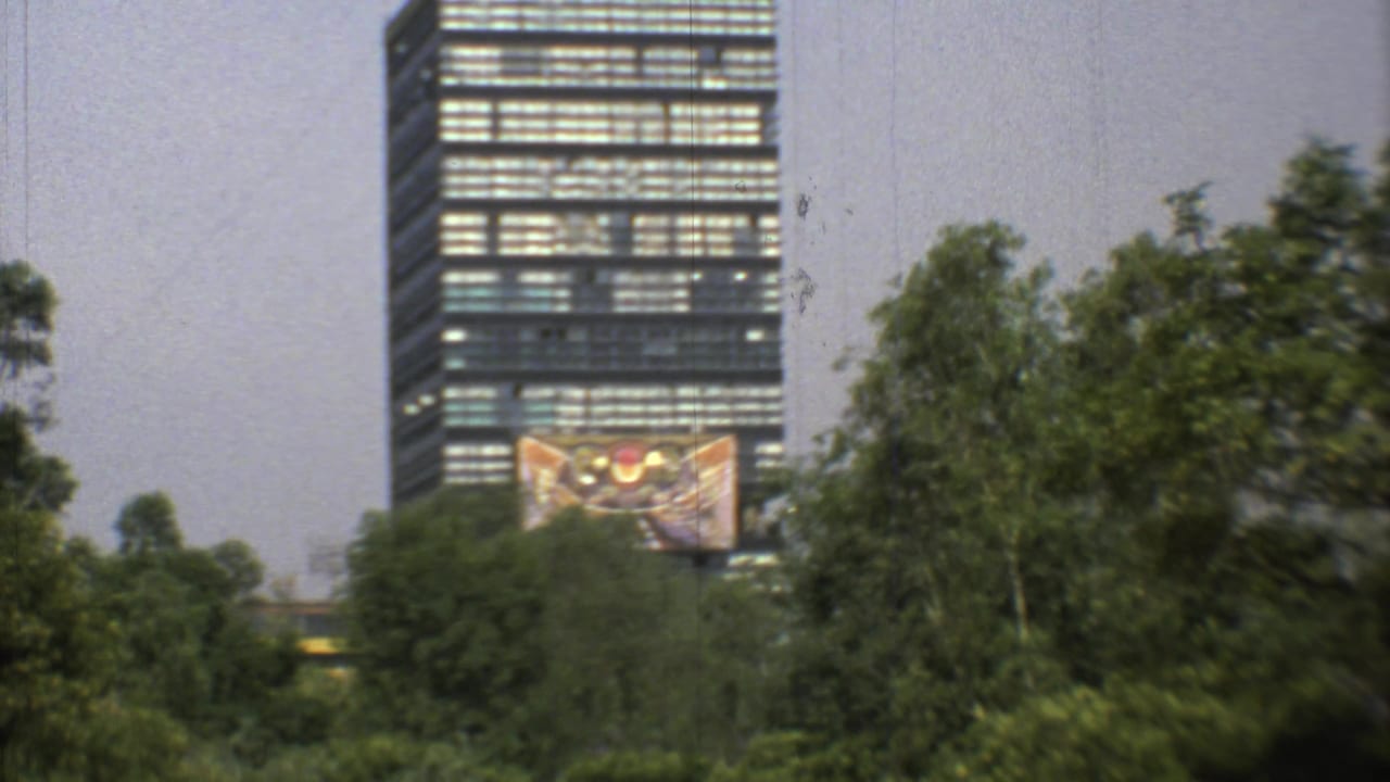 Zoomed 1973 Mexico City view showing a dark modern tower, dense trees, and a large mural or facade artwork near its base.