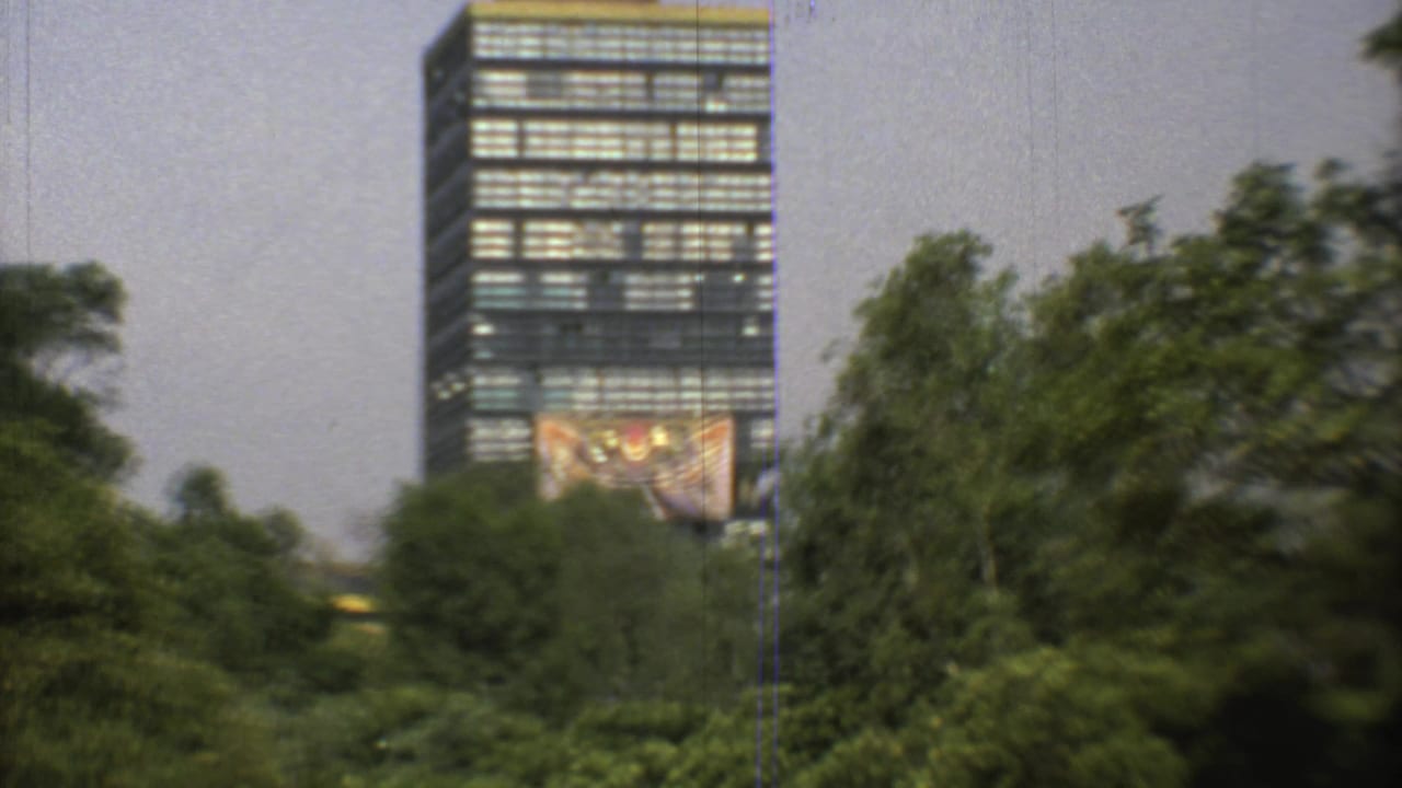 Soft 1973 Mexico City home-movie frame showing a high-rise behind trees, with a large facade artwork still visible near the lower tower.