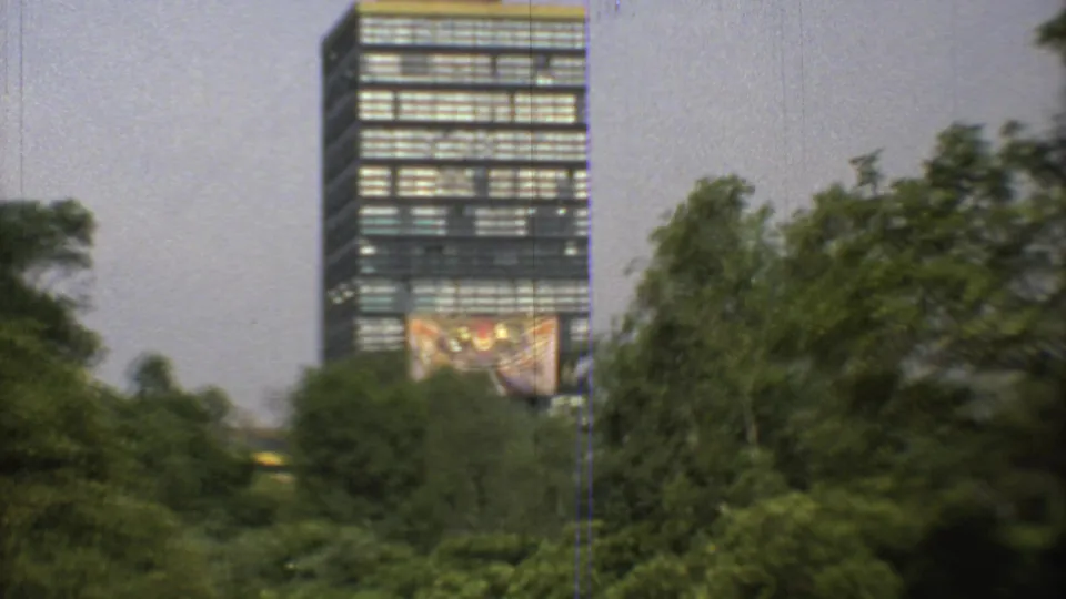 Soft 1973 Mexico City home-movie frame showing a high-rise behind trees, with a large facade artwork still visible near the lower tower.