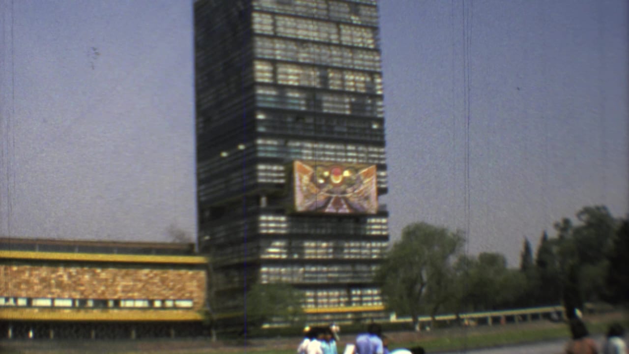 Tall glass building with a large mural, low building edge, trees, and small pedestrians in the foreground.