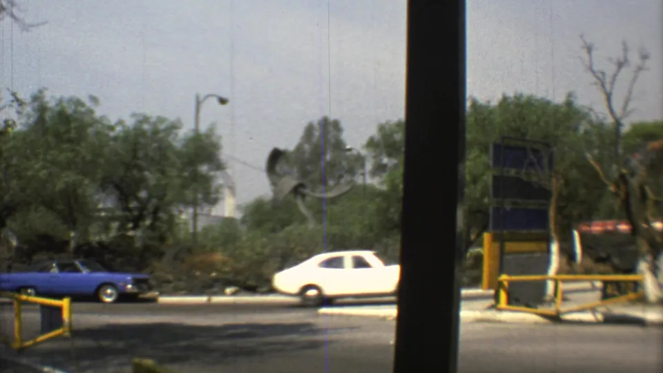 1973 Mexico City 8mm street frame with blue and white cars passing trees, a roadside barrier, and distant sculptural forms.