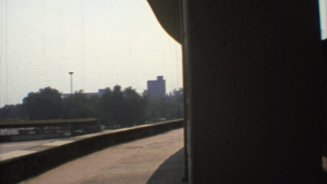 1973 Mexico City 8mm frame looking past a dark structural edge toward a hazy stadium or track area with trees and distant buildings.