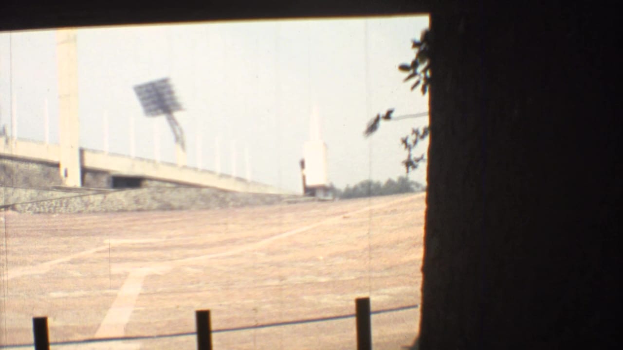 Empty dirt stadium surface with concrete seating, floodlight array, and dark foreground framing.