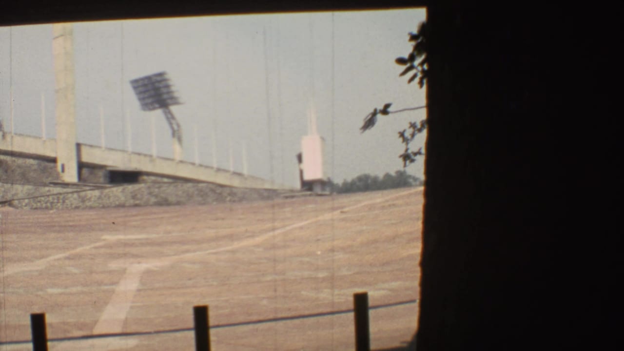 1973 Mexico City 8mm view across an empty dirt stadium surface toward concrete bleachers and a large floodlight array.