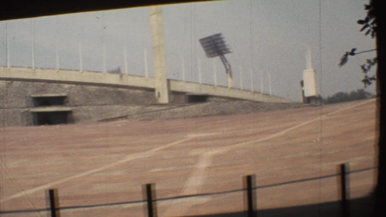 1973 Mexico City 8mm frame showing an empty dirt track, concrete stadium wall, railing posts, and a floodlight tower.