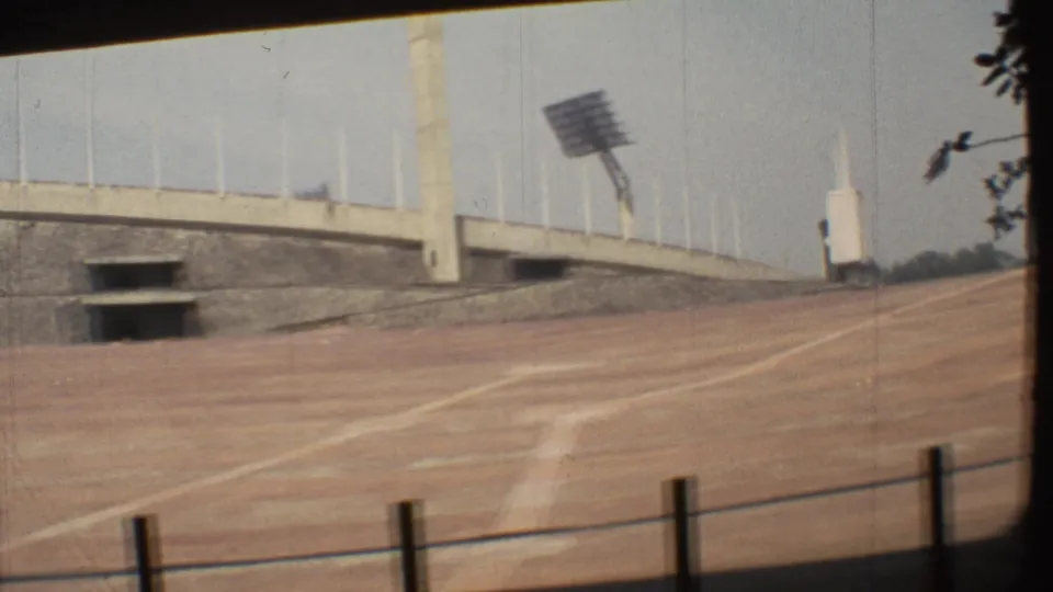 1973 Mexico City 8mm frame showing an empty dirt track, concrete stadium wall, railing posts, and a floodlight tower.