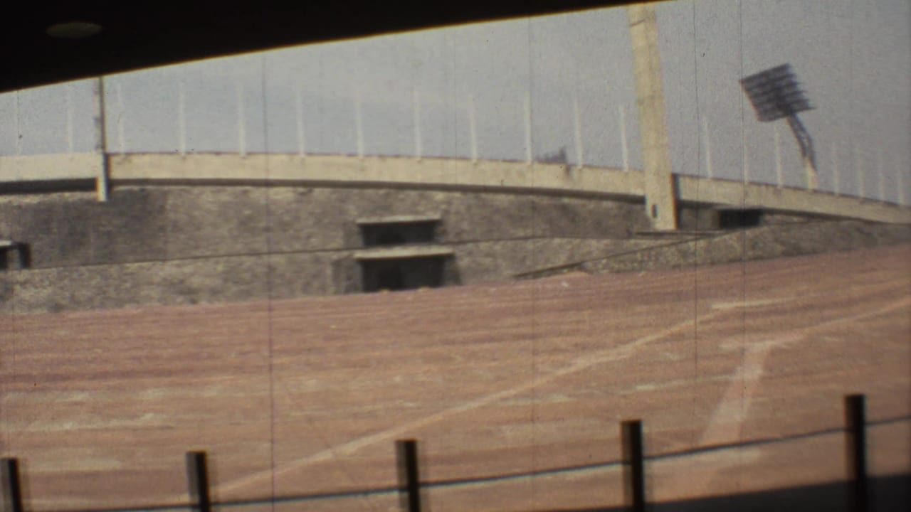 1973 Mexico City stadium view with reddish dirt track, concrete retaining wall, rectangular openings, and a distant floodlight tower.