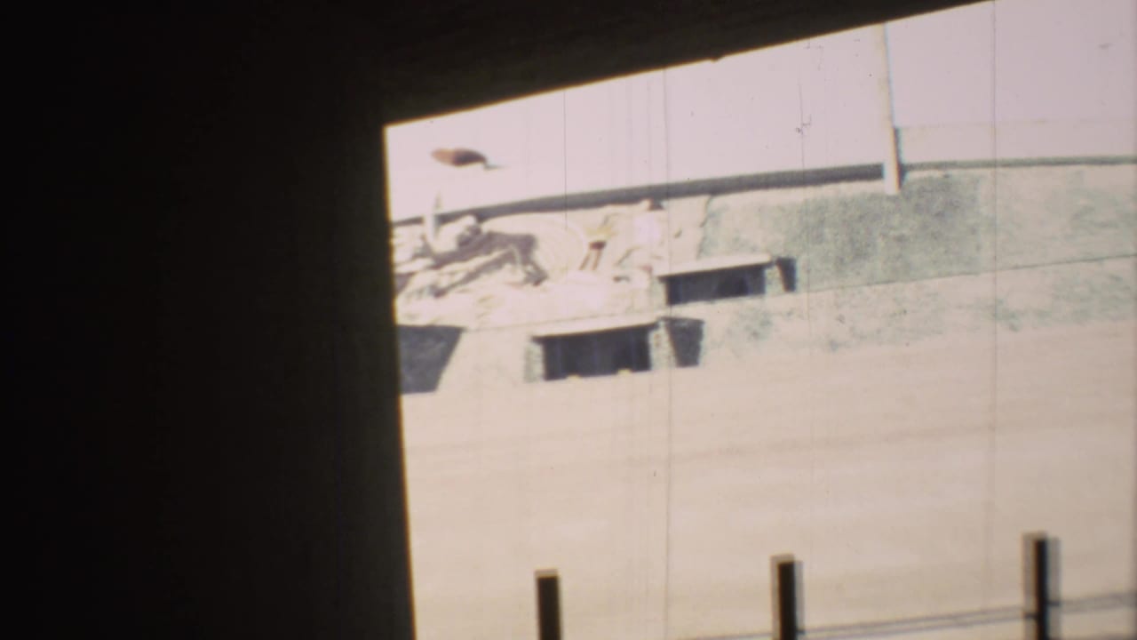 1973 Mexico City street view through a dark edge, showing a pale wall, partial mural forms, small openings, and posts along the sidewalk.