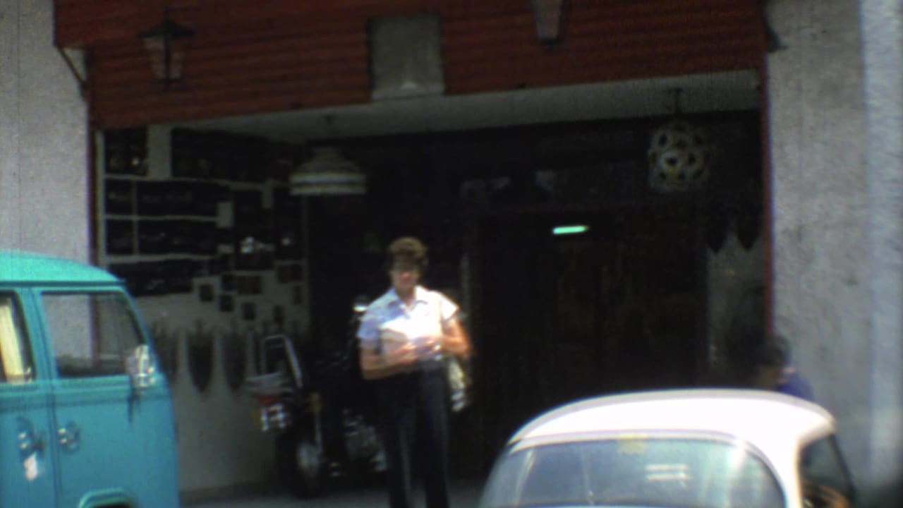 1973 Mexico City street frame showing an adult outside a shaded storefront, with a turquoise van, white car, and motorcycle nearby.
