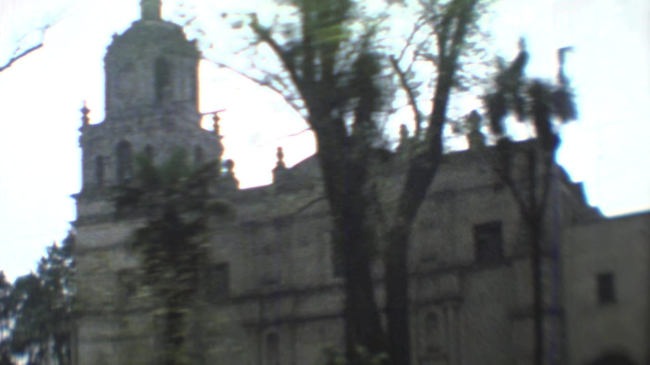 1973 Mexico City 8mm frame showing a weathered church facade and tower seen through trees from a moving camera.