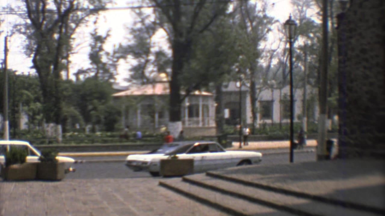 1973 Mexico City home-movie frame showing classic cars along a tree-lined street near steps and a white colonial-style building.
