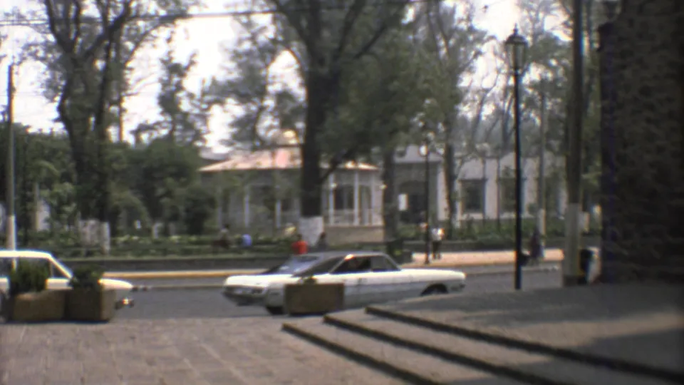 1973 Mexico City home-movie frame showing classic cars along a tree-lined street near steps and a white colonial-style building.