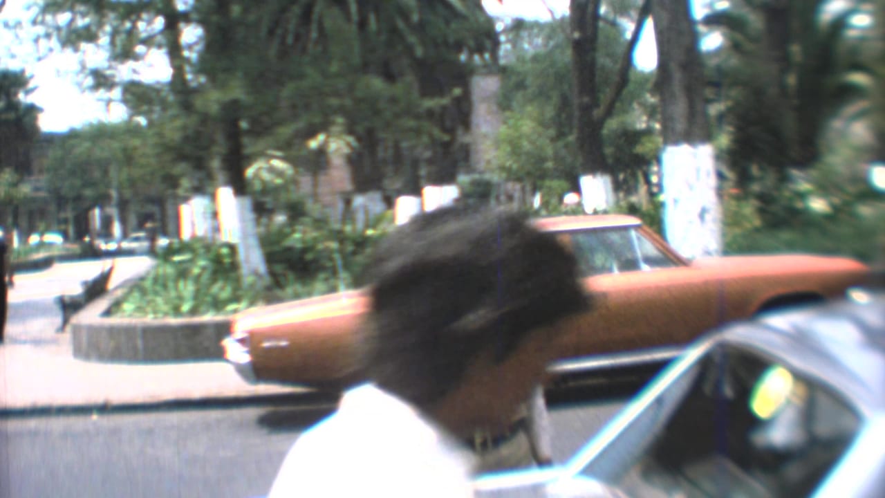 Mexico City street view from 1973 with a blurred pedestrian, orange sedan, parked car roof, and trees in a home-movie frame.