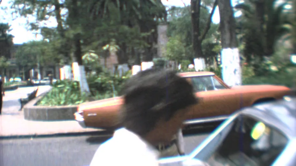 Mexico City street view from 1973 with a blurred pedestrian, orange sedan, parked car roof, and trees in a home-movie frame.
