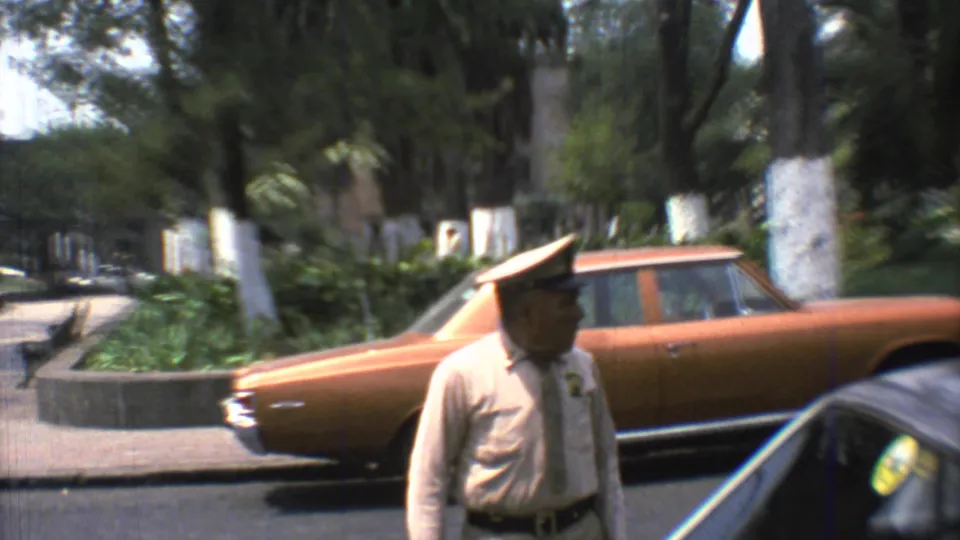 A uniformed police officer stands near an orange sedan on a Mexico City street in 1973, with trees and parked cars behind him.