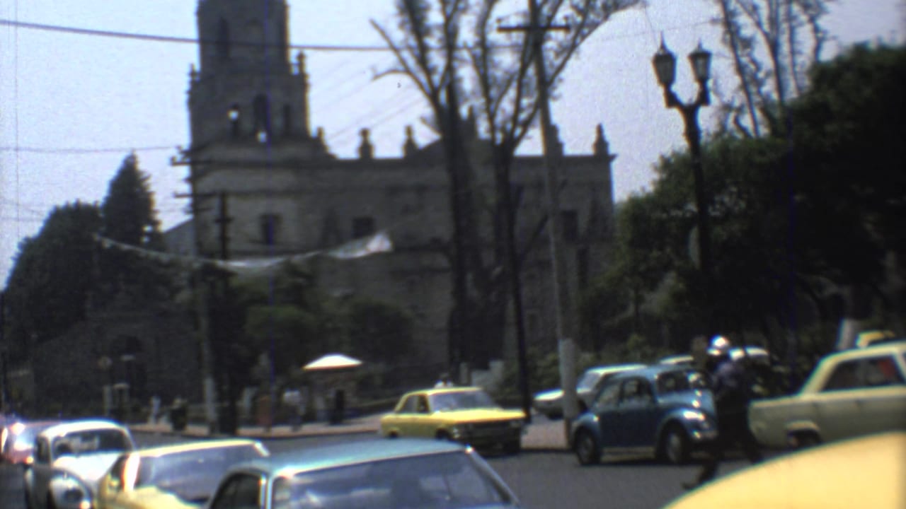 Vintage cars and taxis move through a Mexico City intersection in 1973 with a stone church tower, trees, and streetlamps behind them.