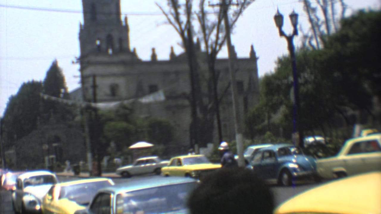 A Mexico City street scene from 1973 shows yellow taxis, a blue Volkswagen Beetle, pedestrians, trees, and a church facade in soft 8mm focus.