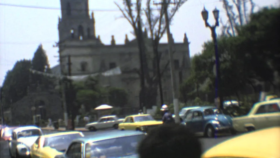 A Mexico City street scene from 1973 shows yellow taxis, a blue Volkswagen Beetle, pedestrians, trees, and a church facade in soft 8mm focus.