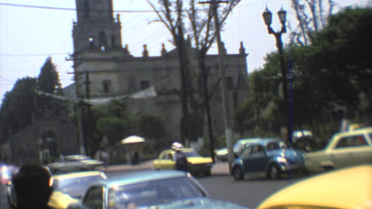 1973 Mexico City street traffic passes a large stone church, with yellow and blue cars, trees, and street lamps in view.