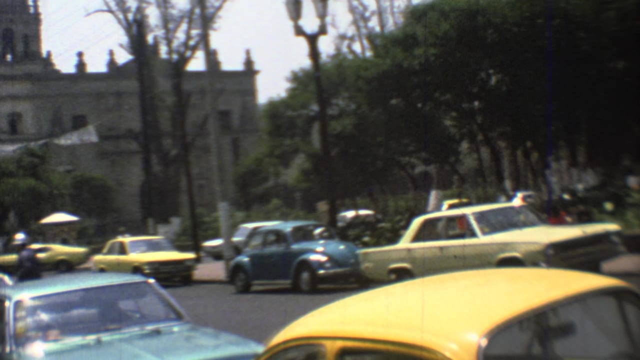 Traffic crosses a tree-lined Mexico City street in 1973, with a church tower and stone facade partly visible behind the cars.