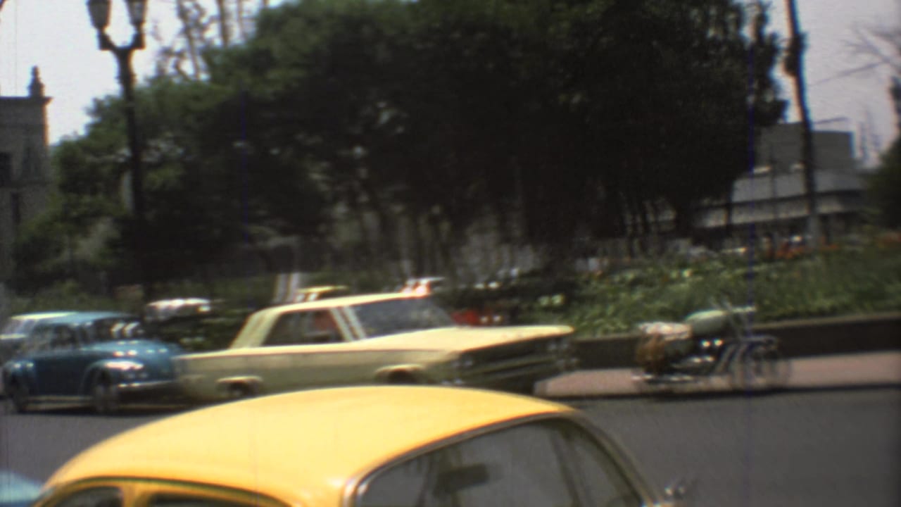 A handheld 8mm view shows 1973 Mexico City traffic beside a landscaped median, with trees and the edge of a church area nearby.