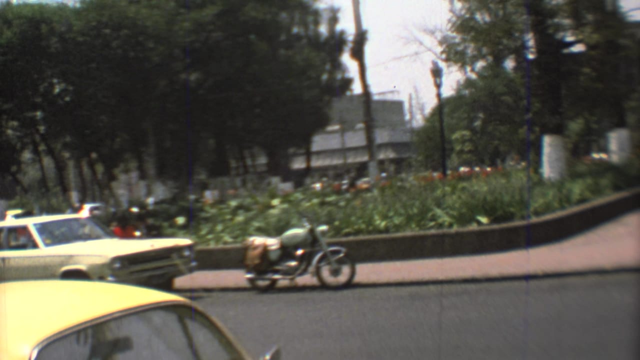A motorcycle and pale car move along a 1973 Mexico City street beside a planted median, trees, and sidewalks.