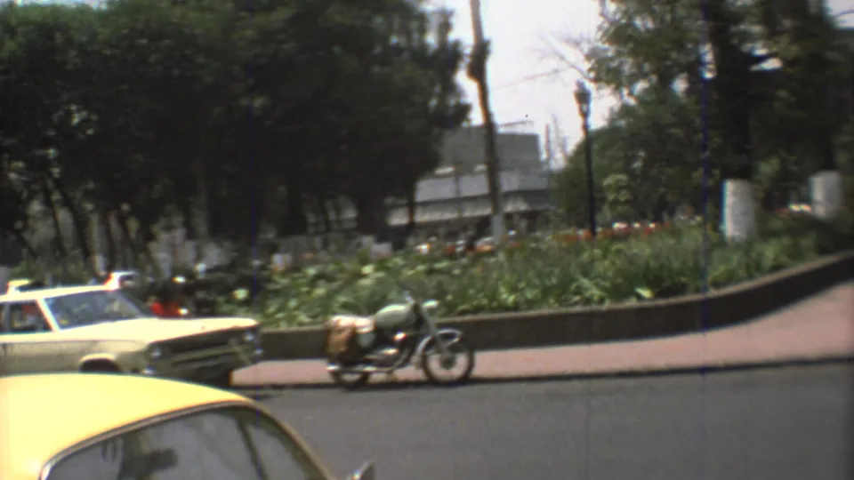 A motorcycle and pale car move along a 1973 Mexico City street beside a planted median, trees, and sidewalks.