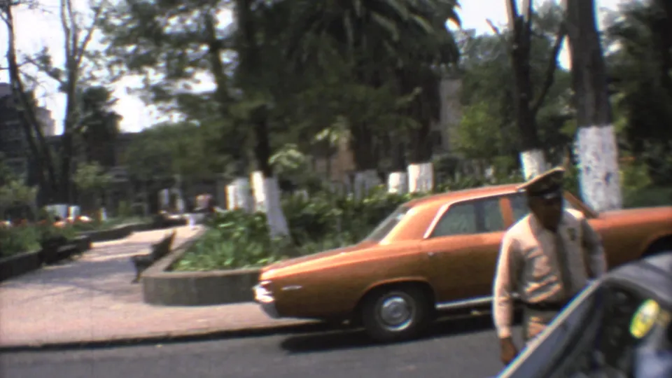 1973 Mexico City street view with a uniformed guard, orange sedan, nearby car, trees, and planted median.