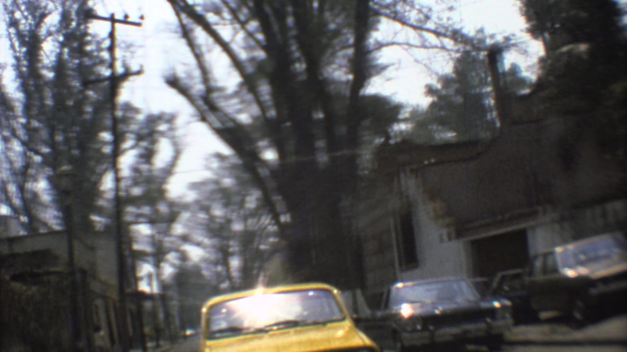 Street view from a moving car with a yellow taxi, parked cars, trees, and a wall.