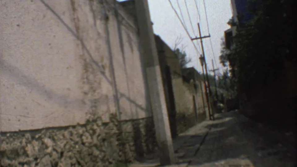 In-car view along a narrow Mexico City street in 1973, showing a long wall, overhead wires, poles, and deep shade.