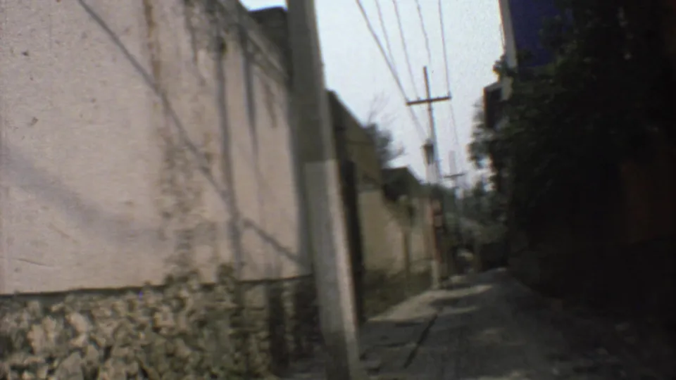 The moving camera passes close to a weathered wall with a stone base on a Mexico City street, with poles and wires ahead.