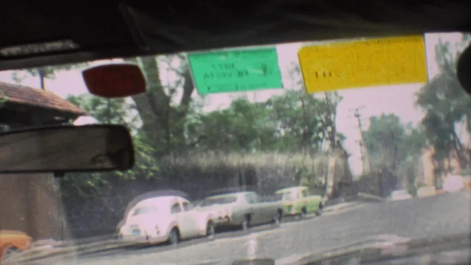 Interior vehicle view in 1973 Mexico City, looking past the rearview mirror and windshield stickers toward parked cars and trees.