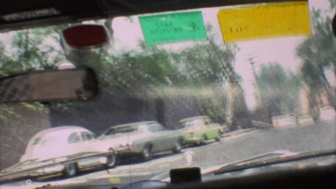 Windshield view of traffic with older cars, palm trees, and inspection stickers visible.