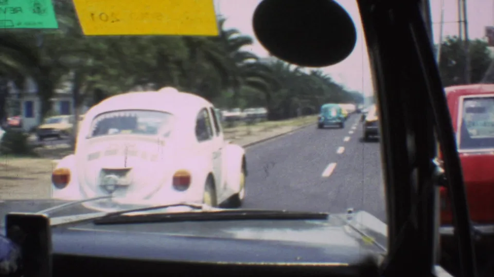 White Volkswagen Beetle moving ahead of the camera car on a Mexico City roadway, with sedans, palms, and windshield stickers visible.