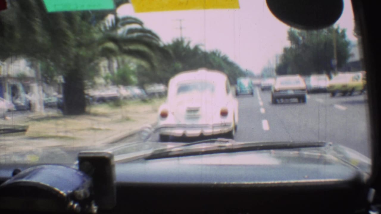 Windshield view of a white Beetle ahead in 1973 Mexico City traffic, with palms, sedans, dashboard, and road markings visible.